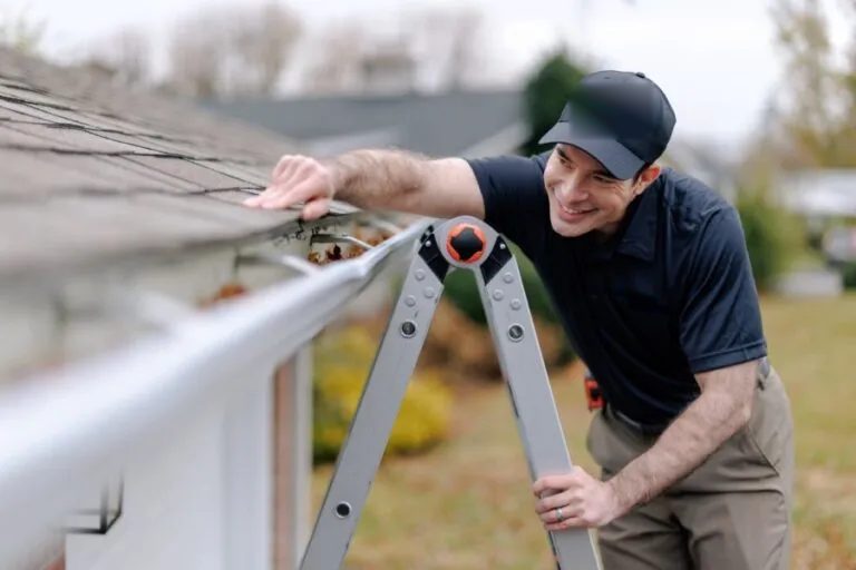 A gutter technician on a ladder checking a gutter full of leaves