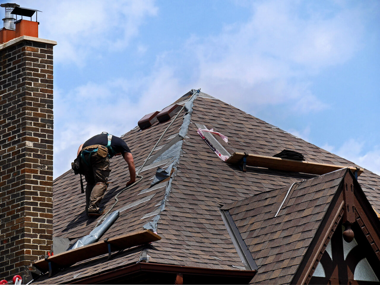 A roofer working on a steep roof section beside a brick chimney